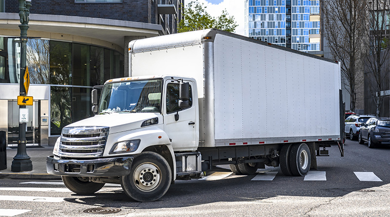 Big rig day cab white semi truck with long box trailer making local commercial delivery at urban city with multilevel residential apartments buildings turning on the city street with crossroad