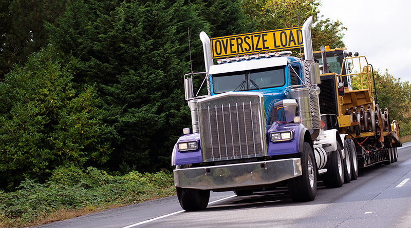 The big powerful rig semi truck with a sign Oversize load on the cab roof and special flat bed trailer which transports a large construction equipment on a straight interstate road